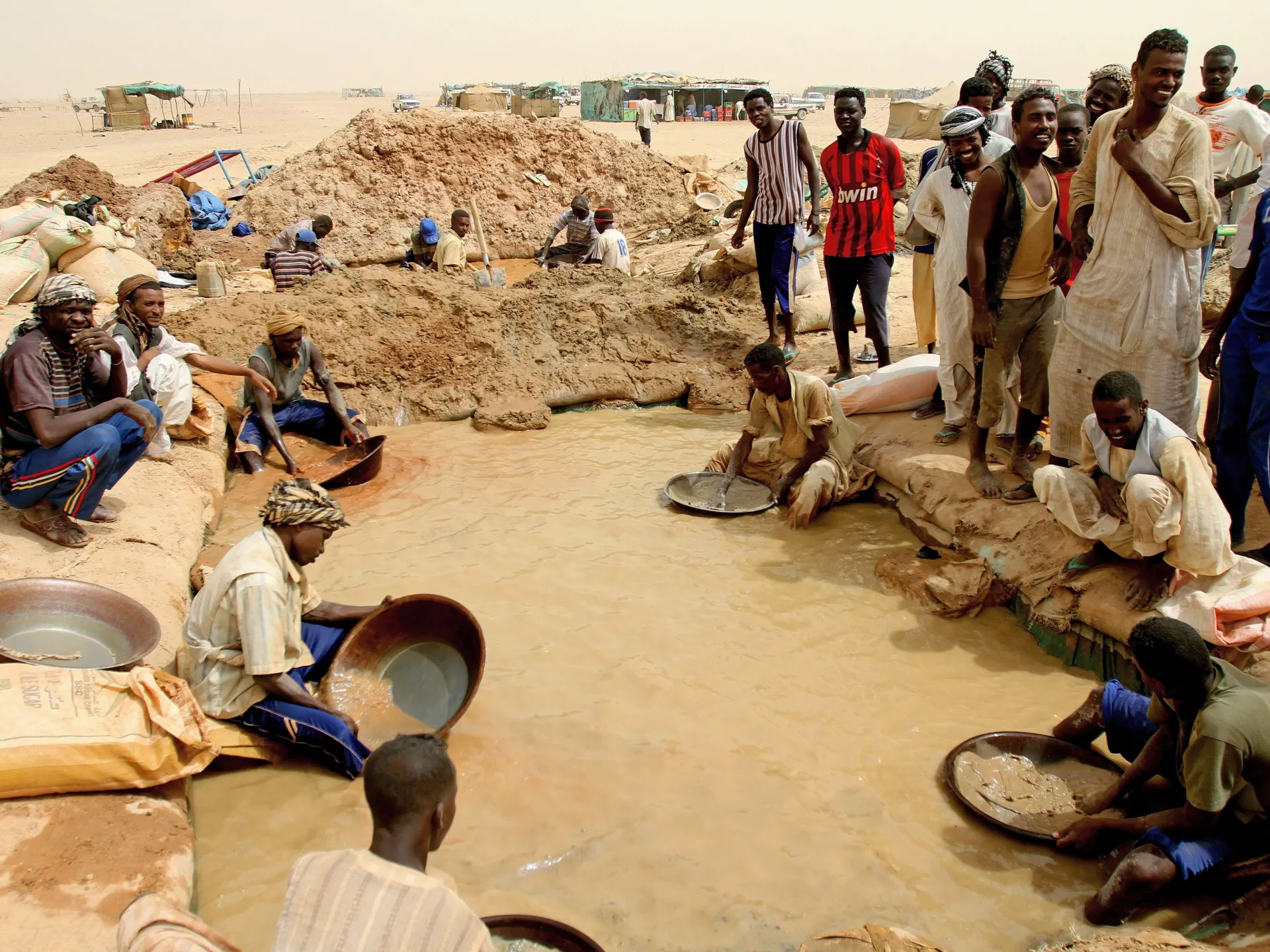 People sift muddy water with pans at an informal mining site in a desert landscape.