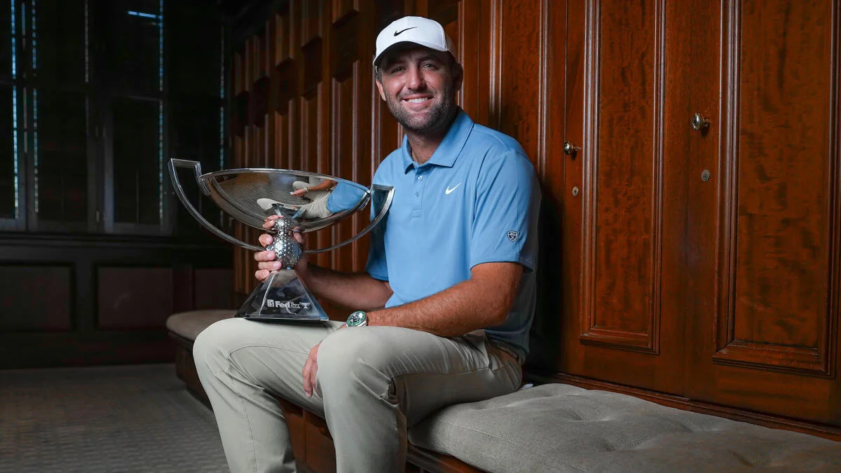 Smiling golfer in blue polo and white cap holds a large silver trophy while seated indoors.