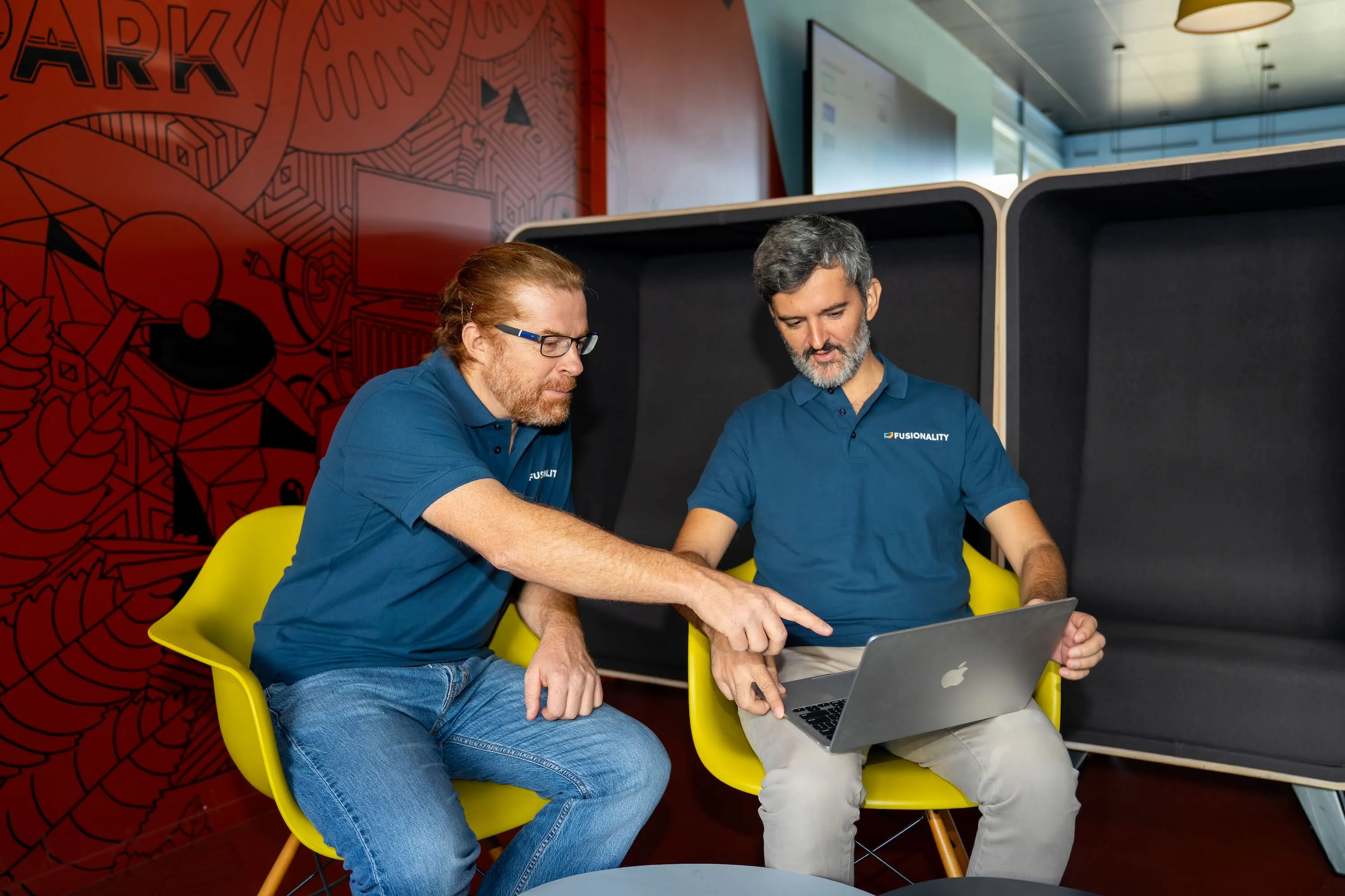 Two men in Fusionality shirts review a laptop together in a colorful office workspace.