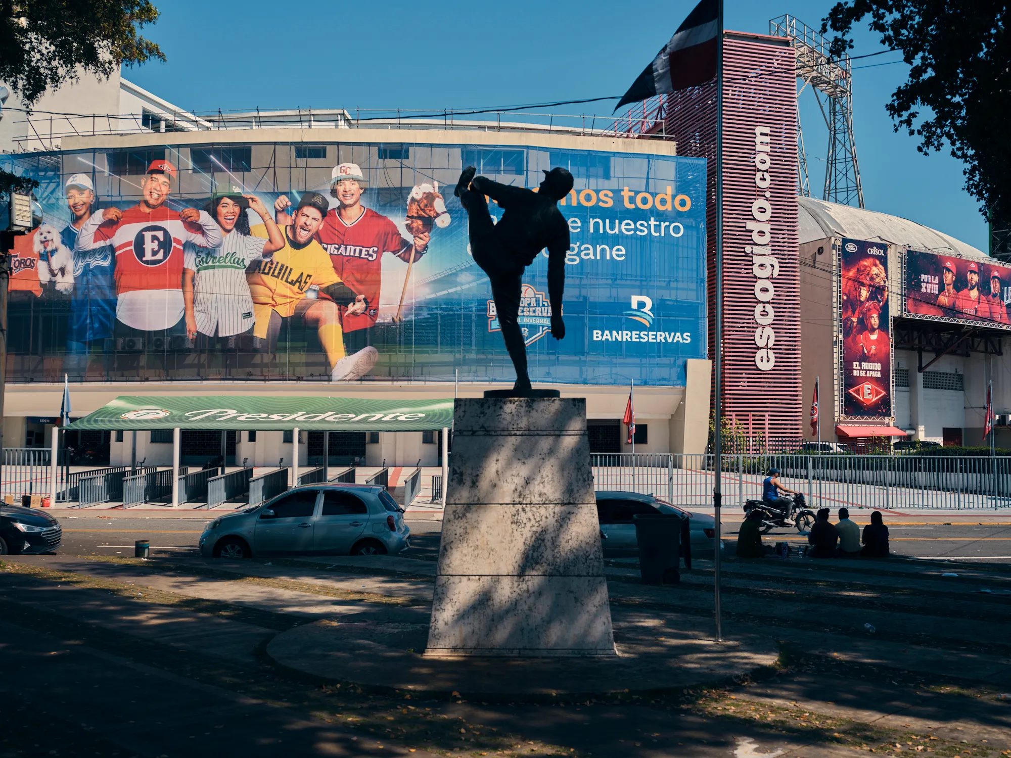 Baseball statue stands before a stadium covered with colorful Dominican baseball advertisements.