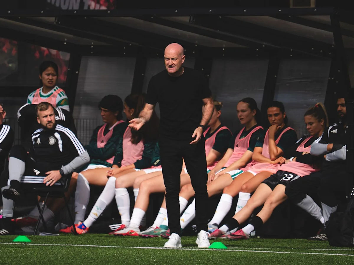 A bald football coach gestures in front of seated players on a team bench.