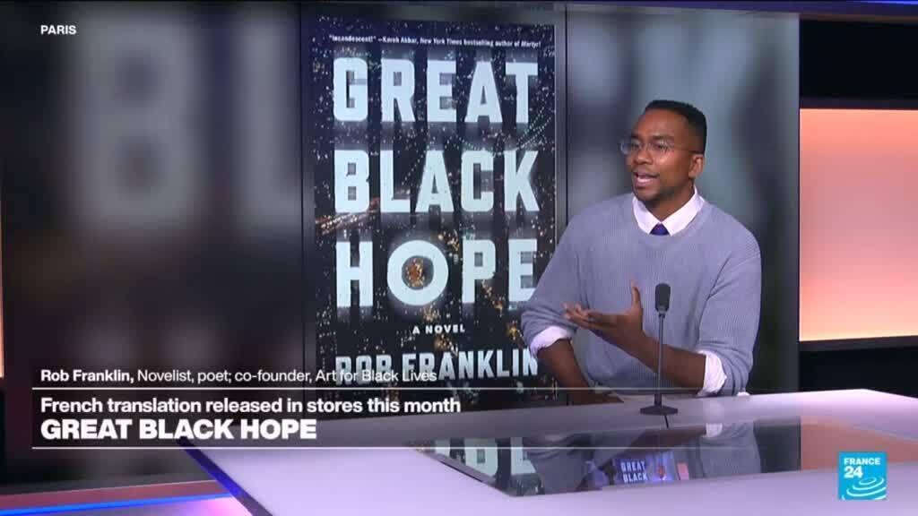 A man in a lavender sweater speaks beside a large Great Black Hope book cover in a television studio.