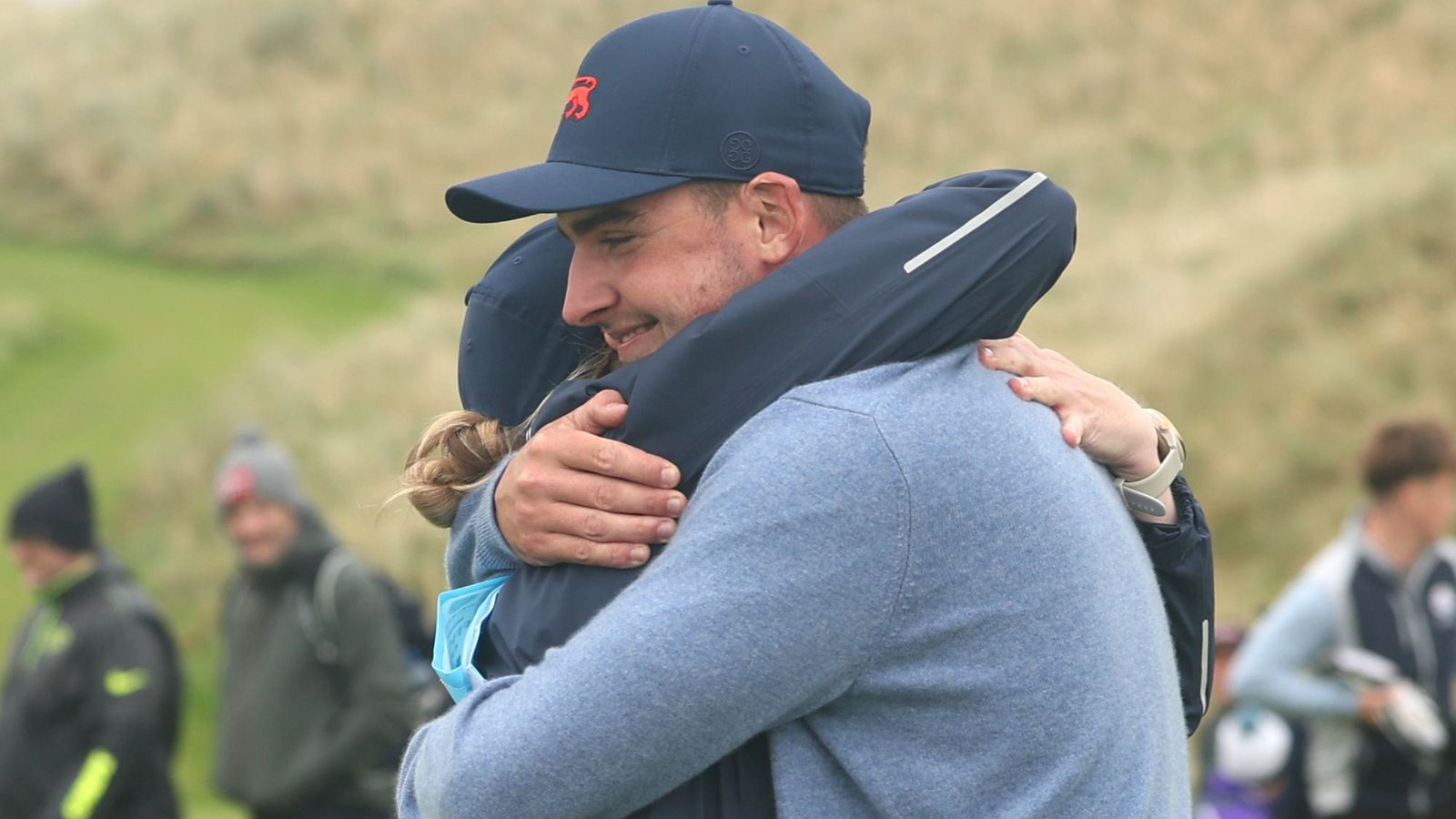 Two people embrace outdoors, one wearing a navy cap and the other a blue-gray sweater.