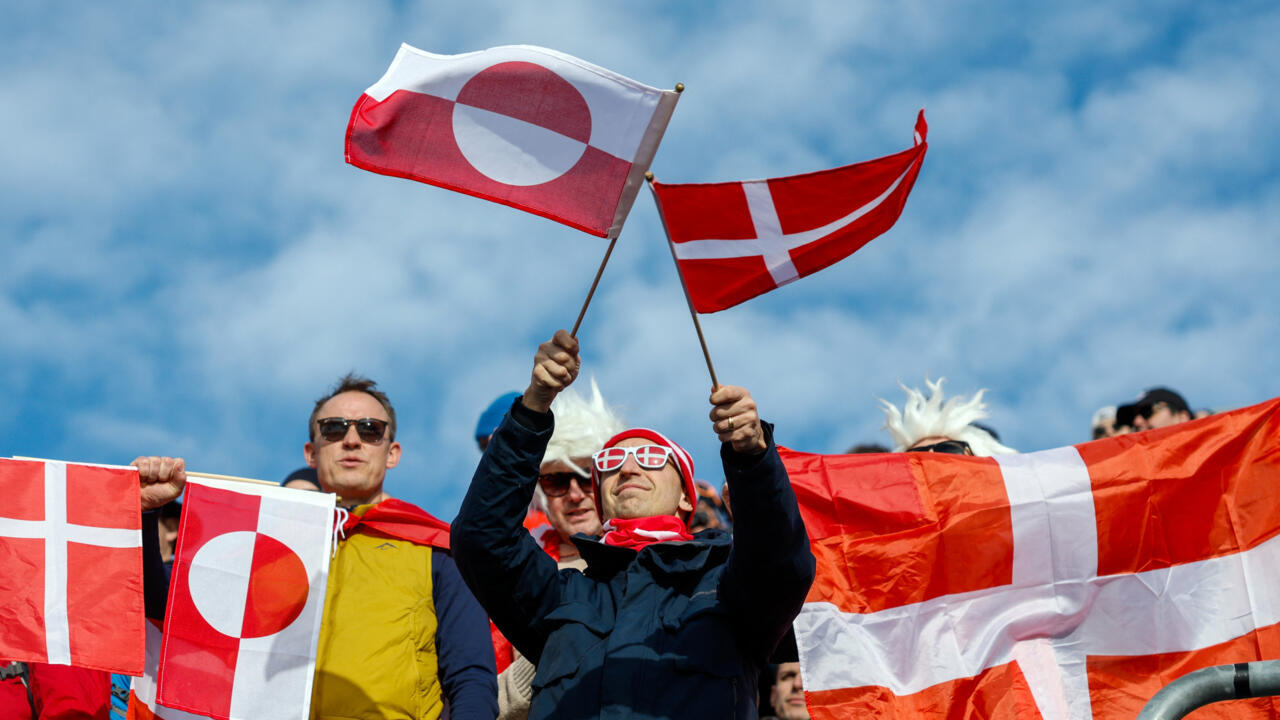 Crowd members wave Greenlandic and Danish flags beneath a bright blue sky.