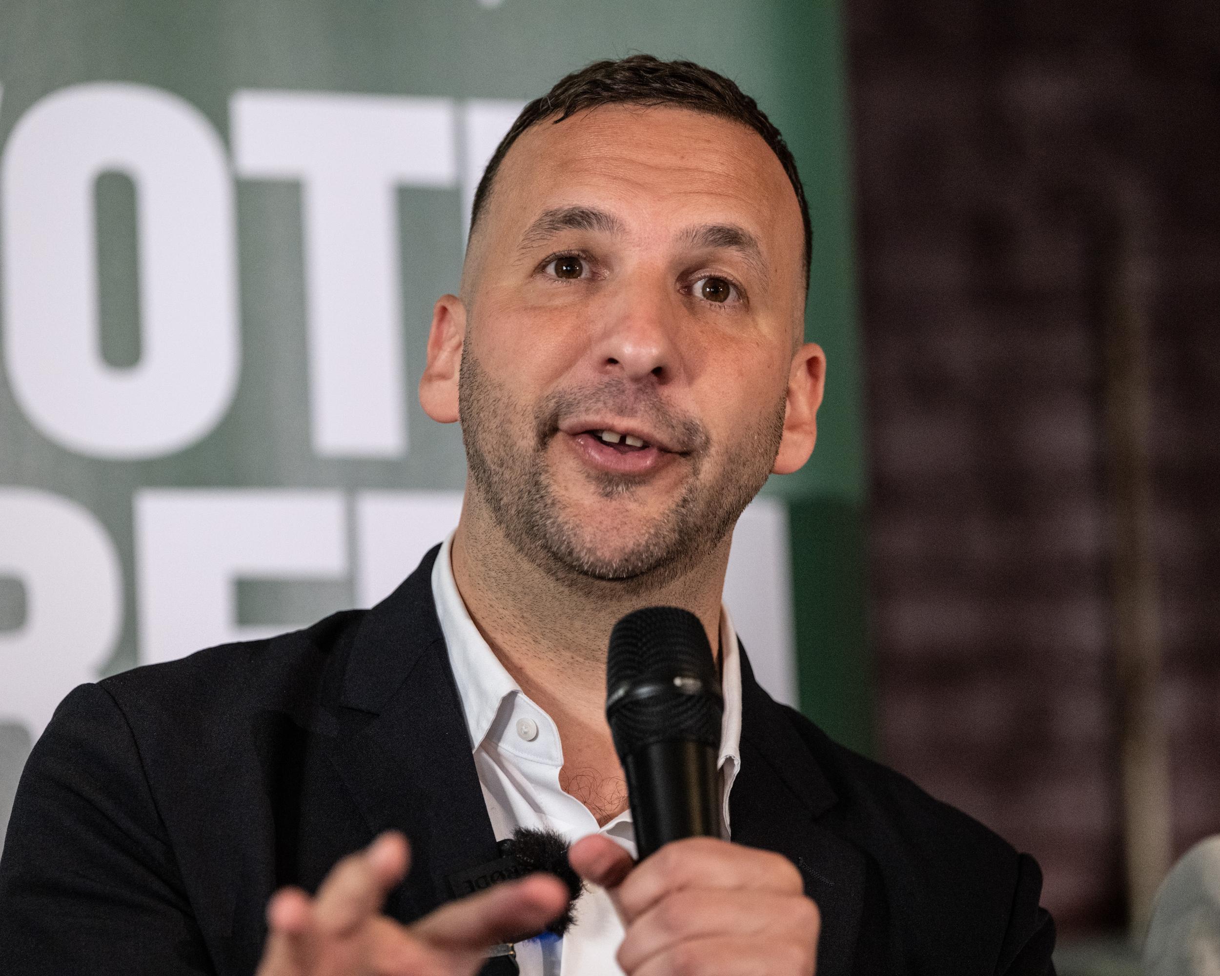 Man in dark suit speaking into a microphone during a public event