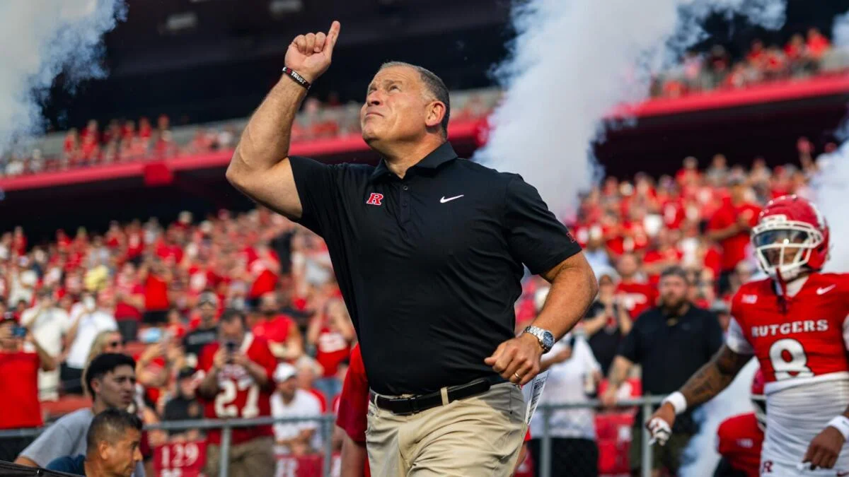 Rutgers coach in black polo points upward while a football player stands nearby on the sideline