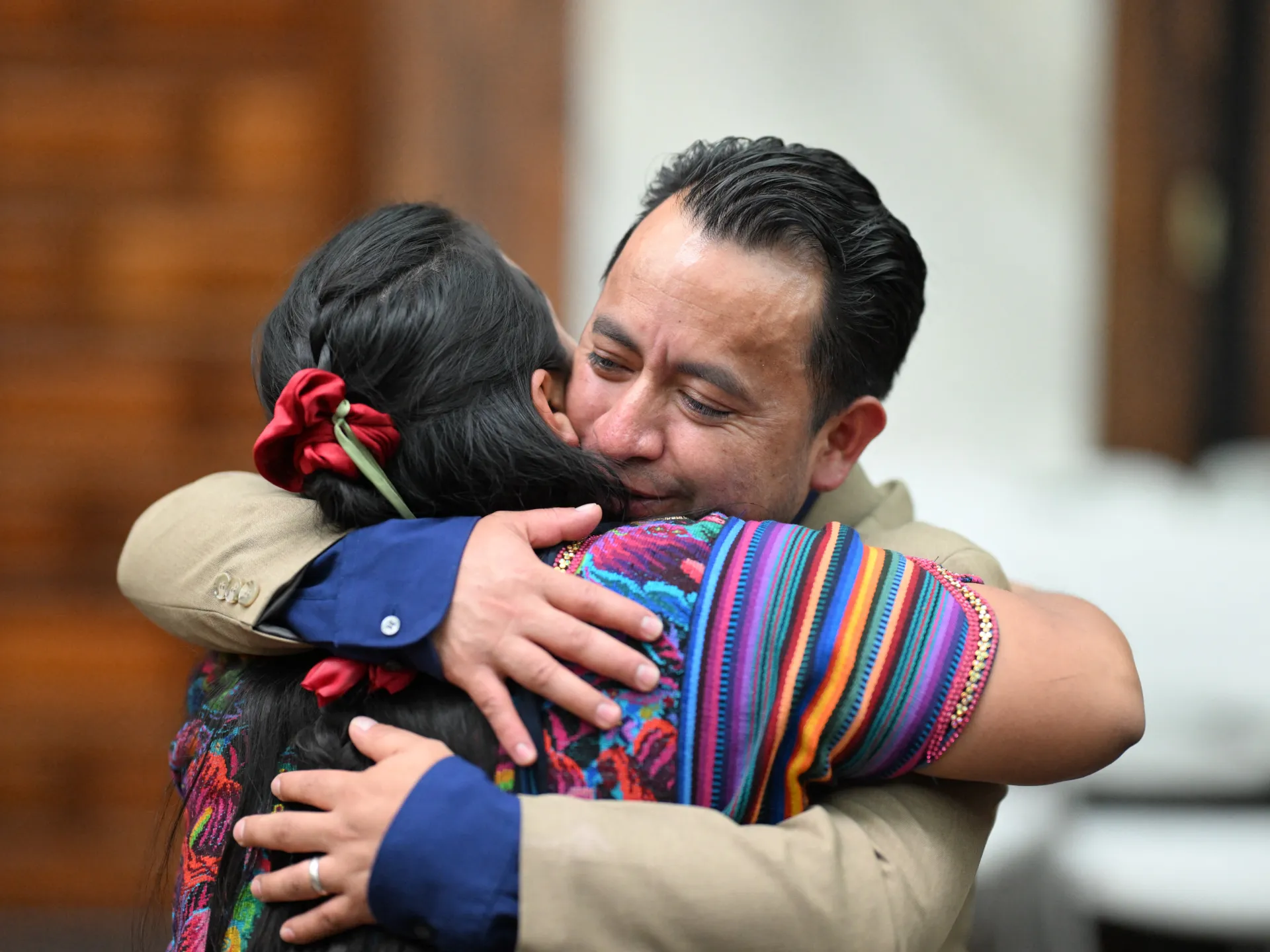 A man embraces a woman wearing colorful traditional clothing, both shown in close-up.