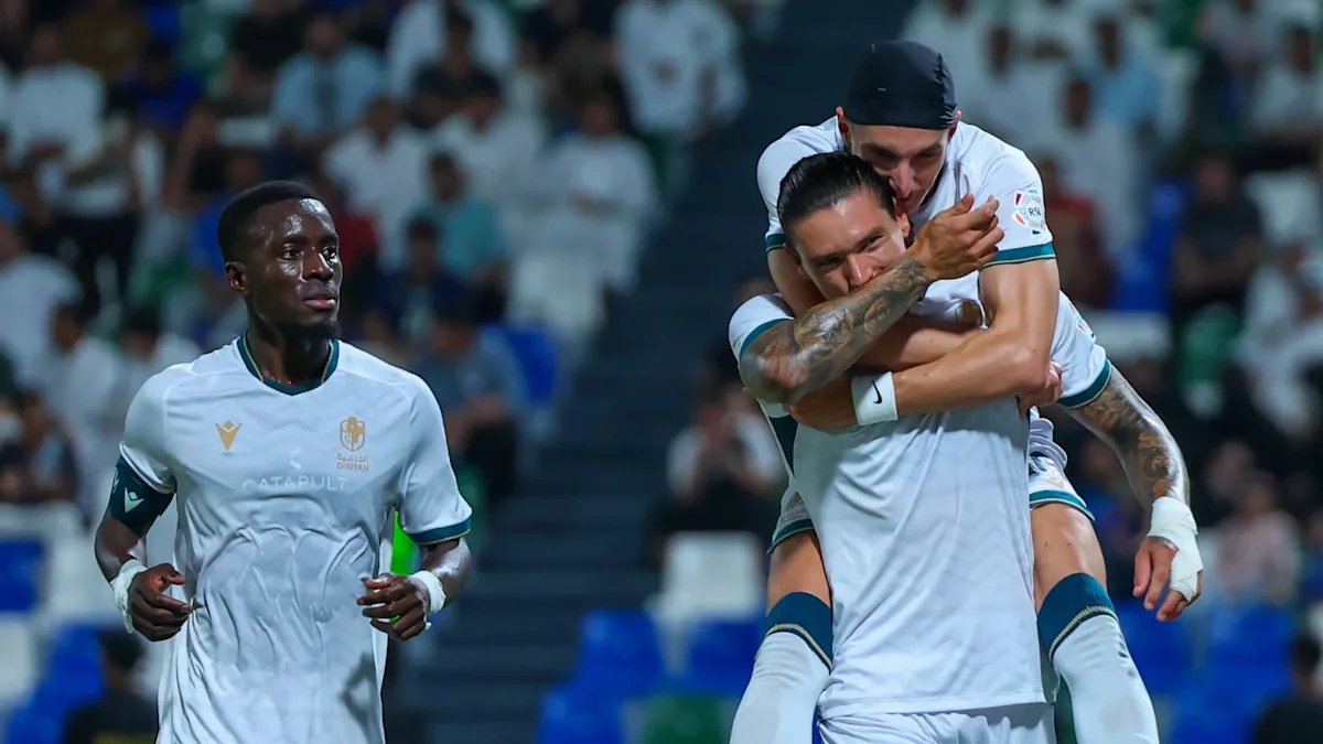 Three footballers in white kits celebrate together during a match, with one player carrying another.