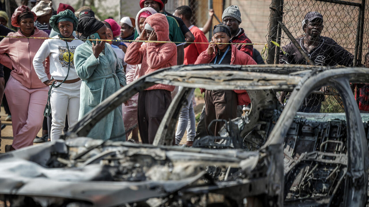 People stand behind a cordon near a burned-out vehicle, with a chain-link fence in the background.