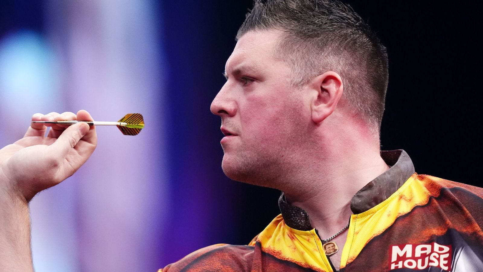 Darts player raises a dart toward his face during a brightly lit televised match.