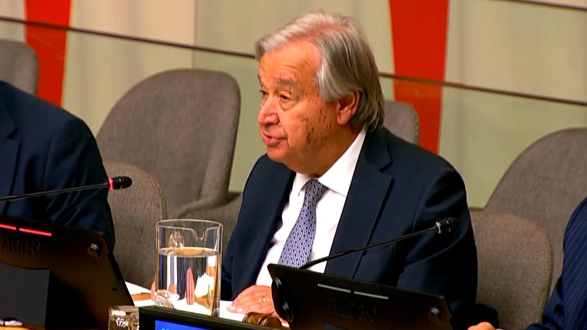 Gray-haired man in dark suit and patterned tie speaking at a conference table with microphones