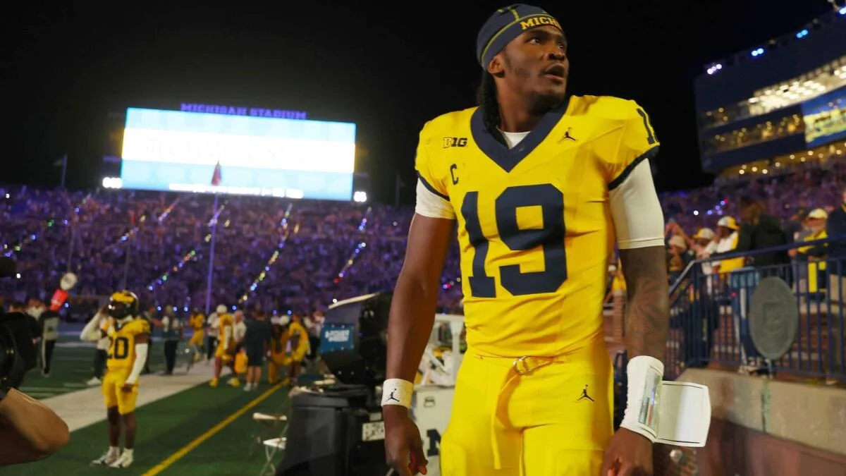 Football player in Michigan uniform walking past the sideline during a night game.