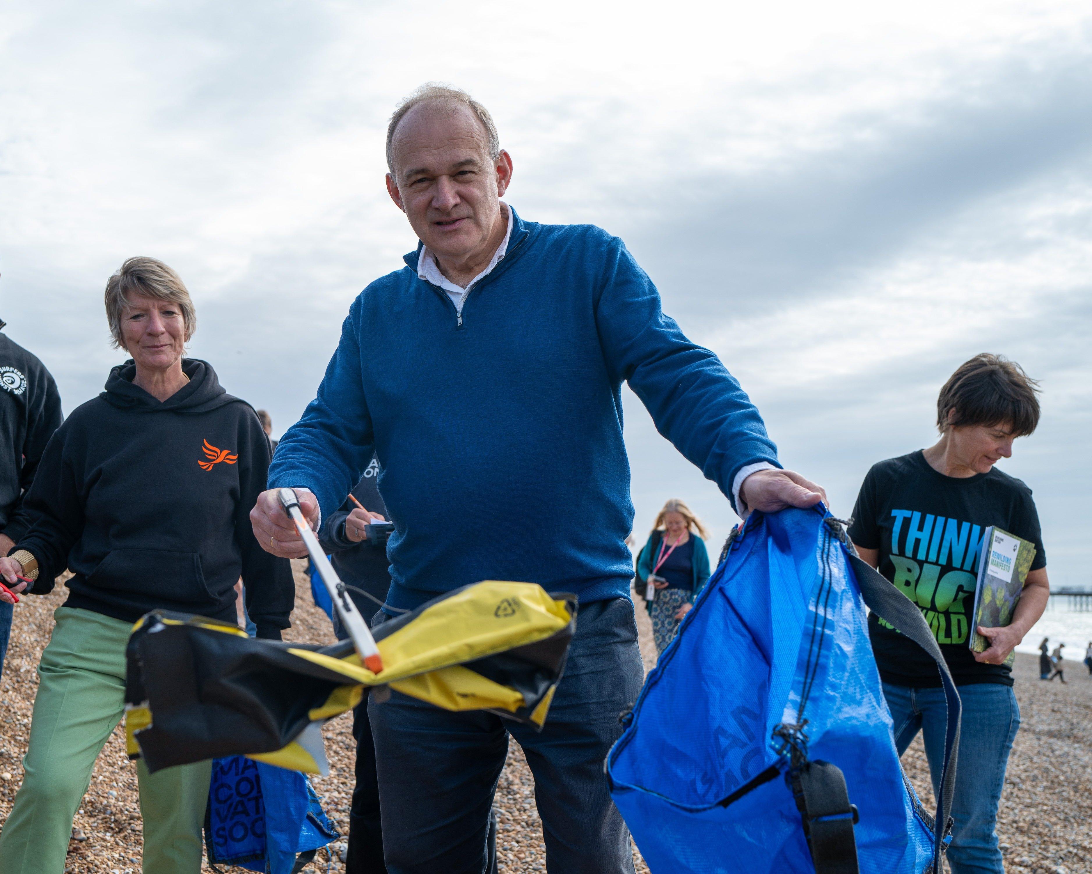 People collect rubbish in large bags on a pebble beach under a cloudy sky.
