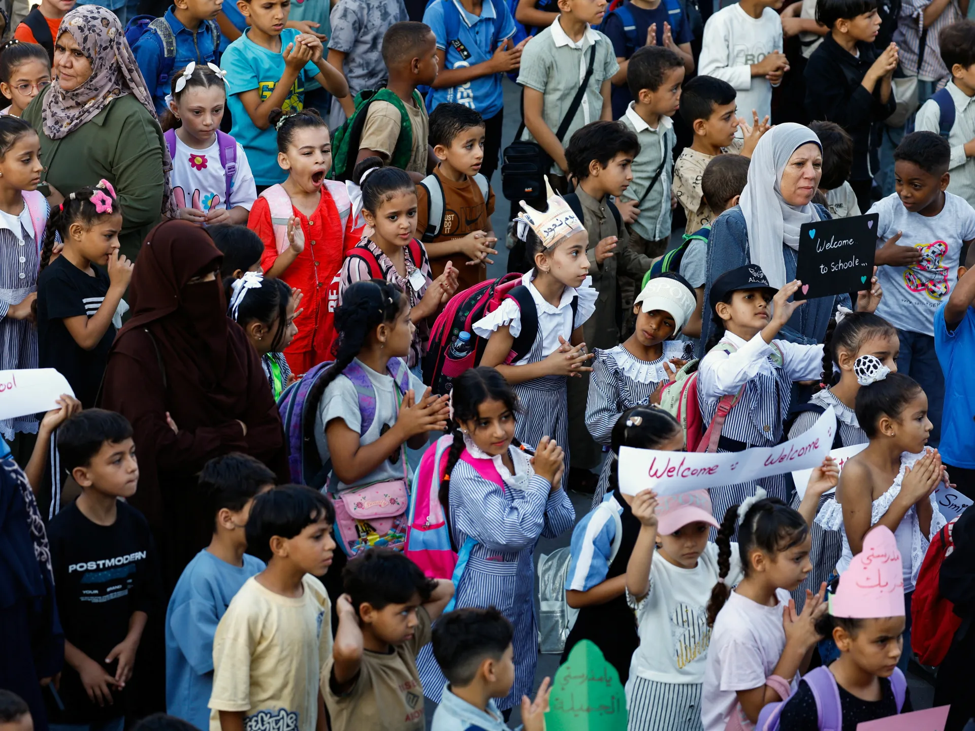 A crowd of children and adults gathers, with several children holding welcome signs and wearing school uniforms.