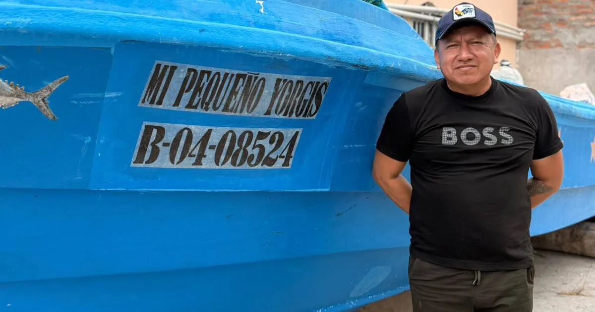 Man in black shirt and cap standing beside a blue fishing boat with painted lettering.