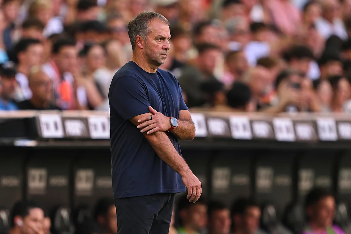 Football coach in navy shirt stands with arms crossed beside the pitch during a match