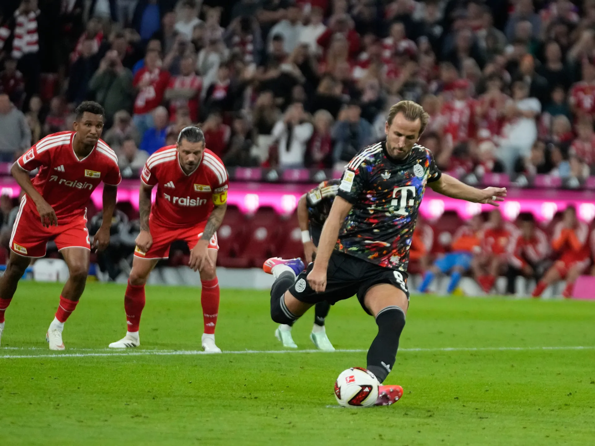 A player in a dark patterned kit strikes a ball as two red-shirted players watch nearby.