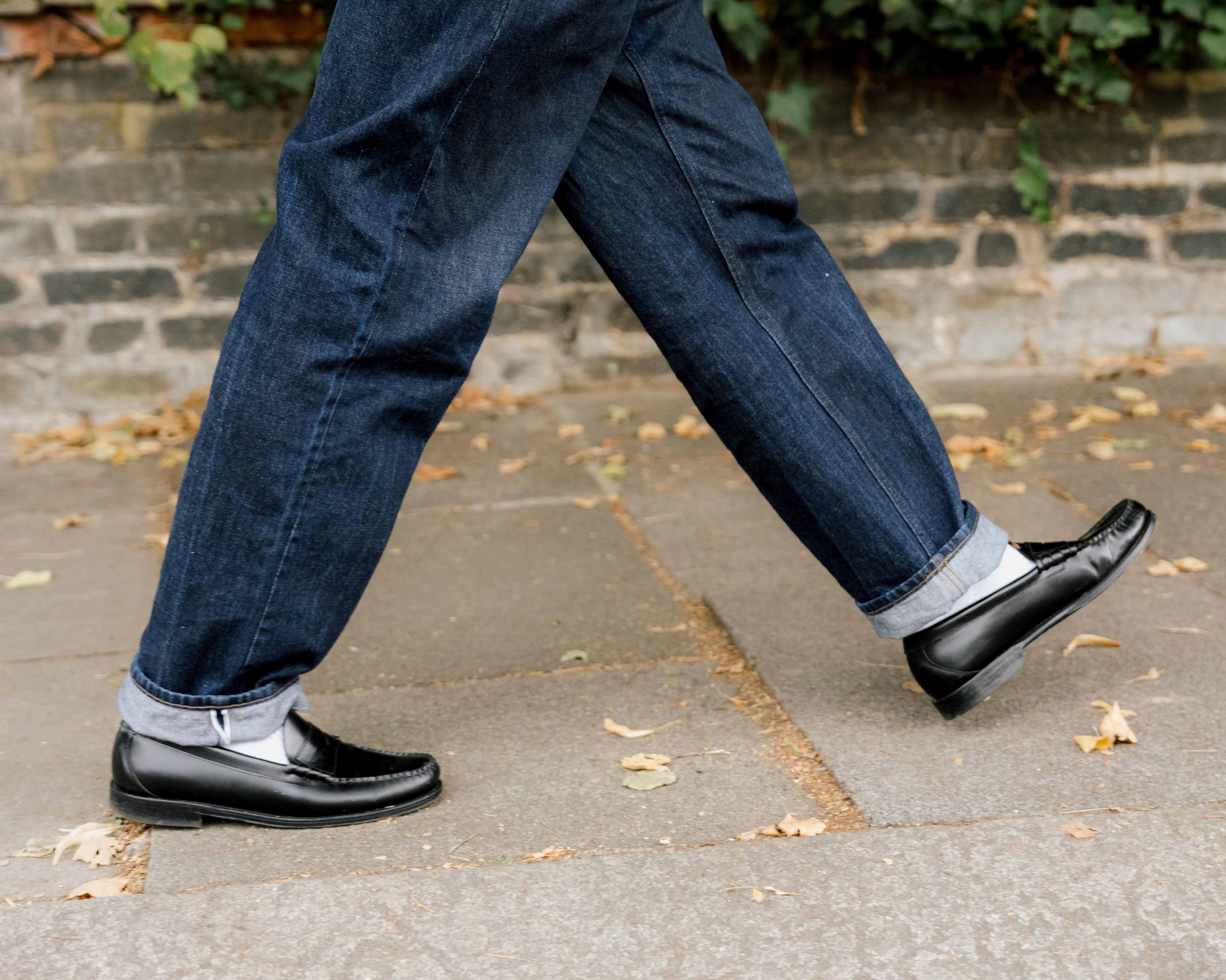 Close-up of a person walking outdoors in cuffed dark jeans, white socks, and black loafers.