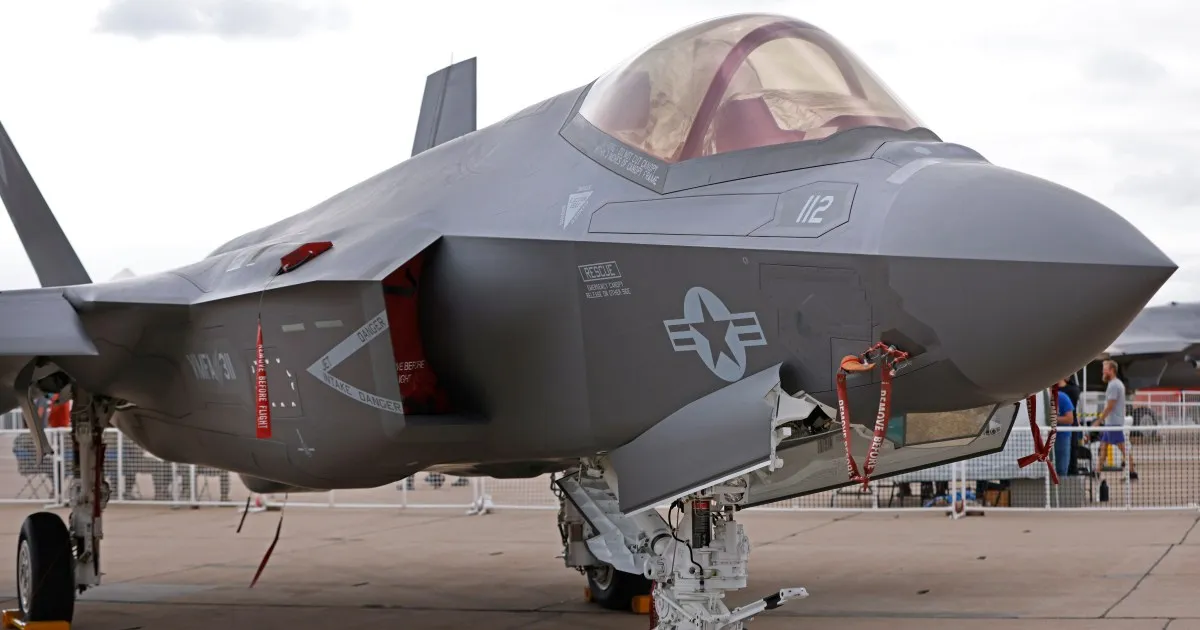 A gray stealth fighter jet parked on an airport apron, with ground equipment and people behind it.