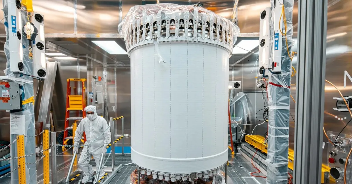 Large cylindrical scientific apparatus surrounded by equipment and a researcher in protective clothing inside a cleanroom.