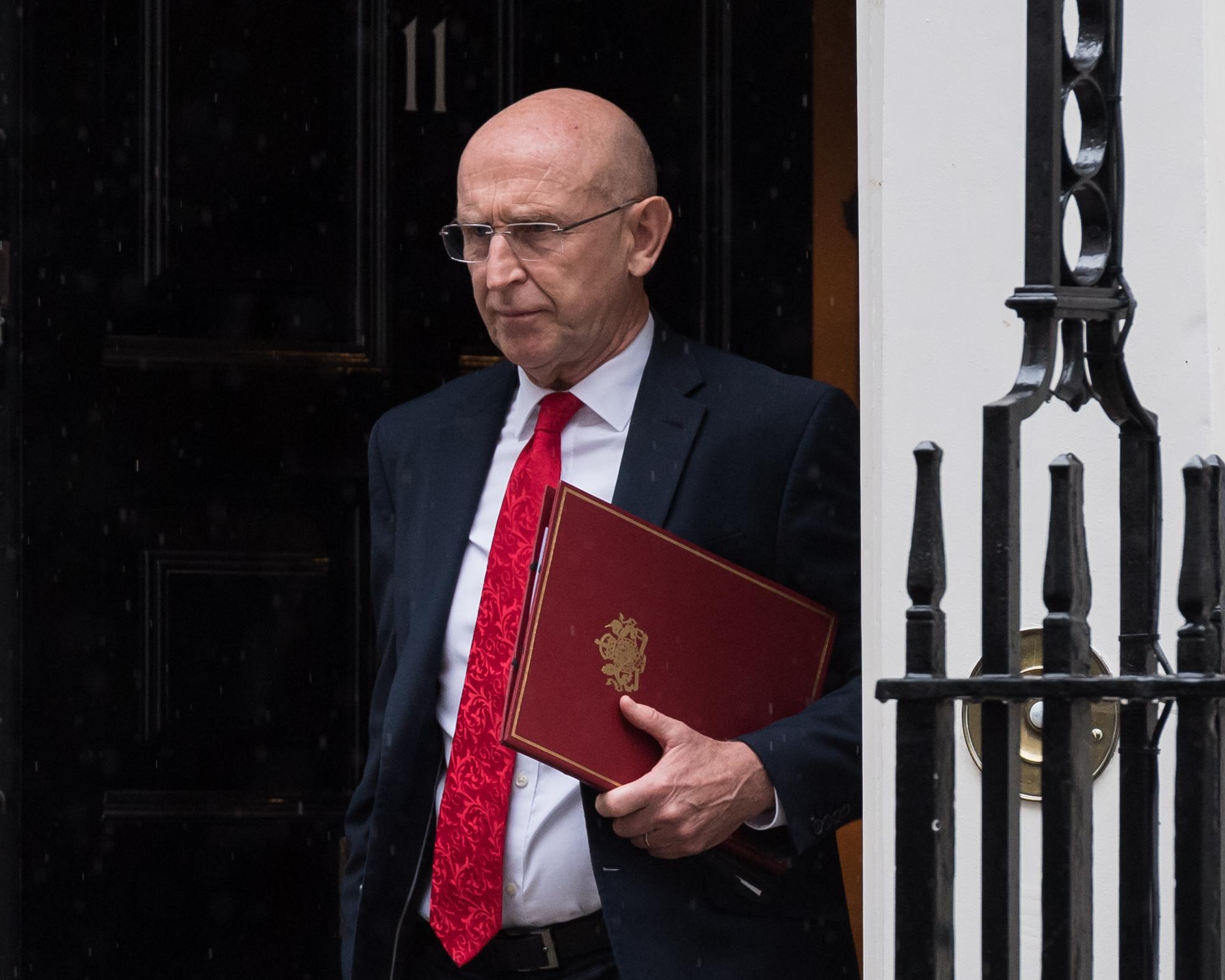 Bald man in glasses holds a red folder beside a large ceremonial chair.