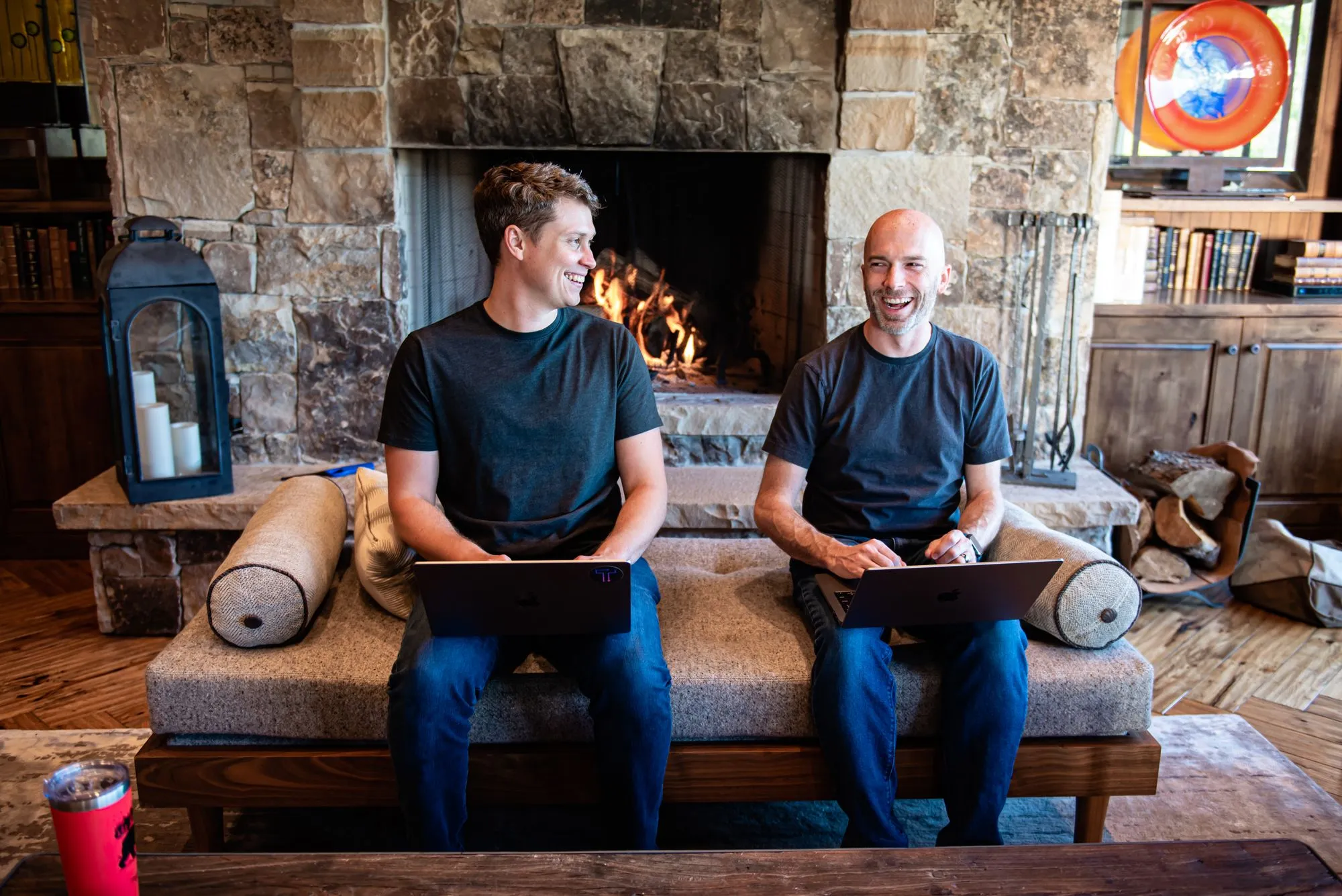 Two men smiling with laptops while seated before a stone fireplace in a rustic living room