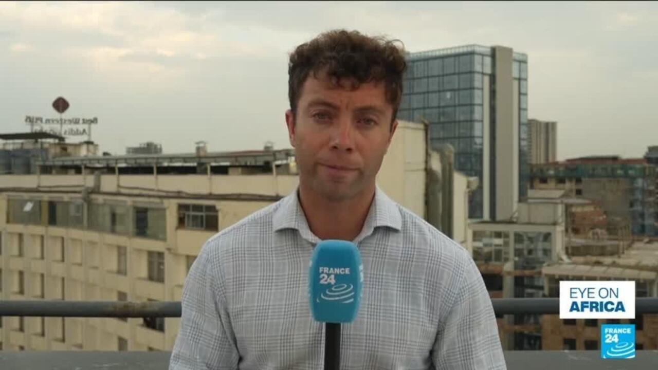 A male reporter holds a France 24 microphone on a rooftop, with city buildings behind him.