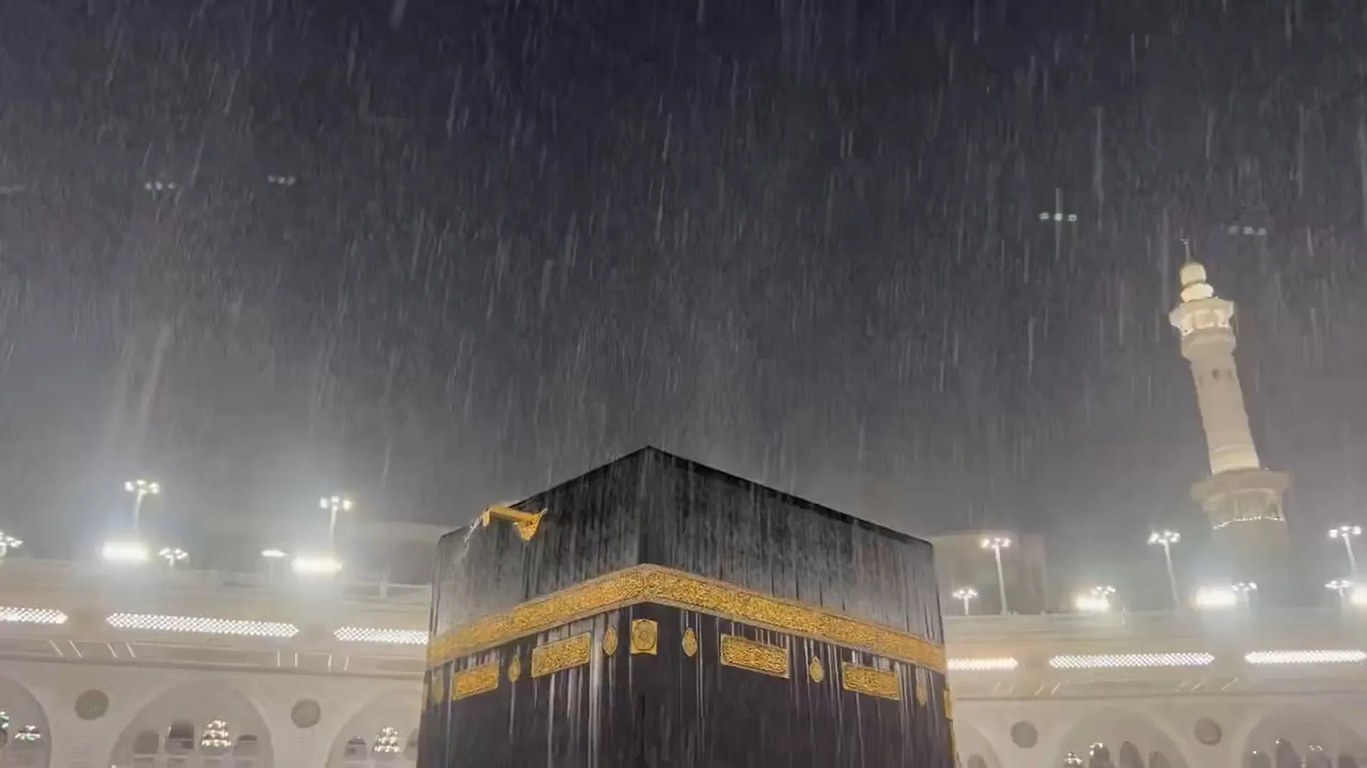 Heavy rain falls around the Kaaba, with mosque lights and a minaret visible through mist.