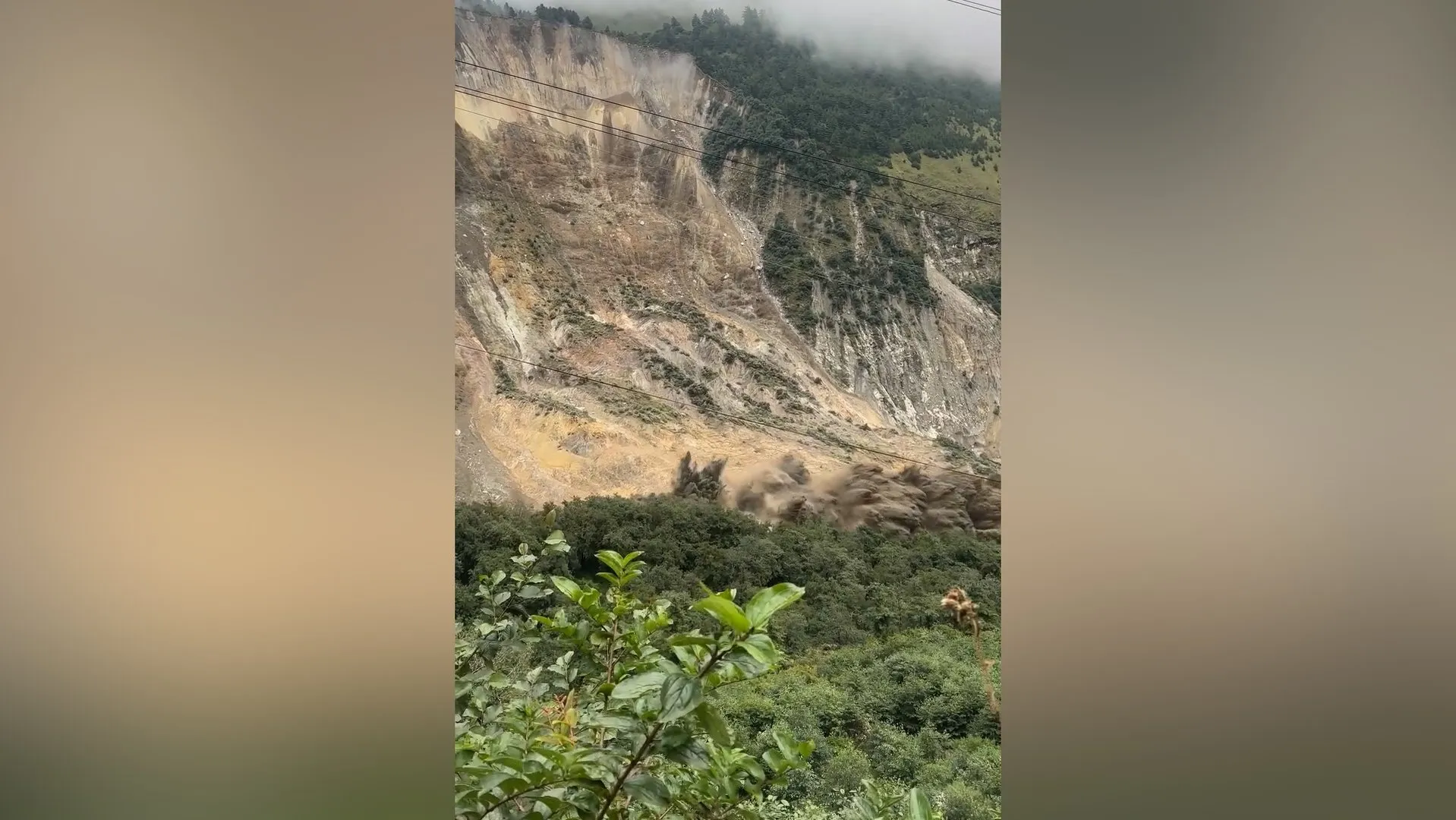A large landslide sends dust and debris down a steep mountainside above dense green forest.