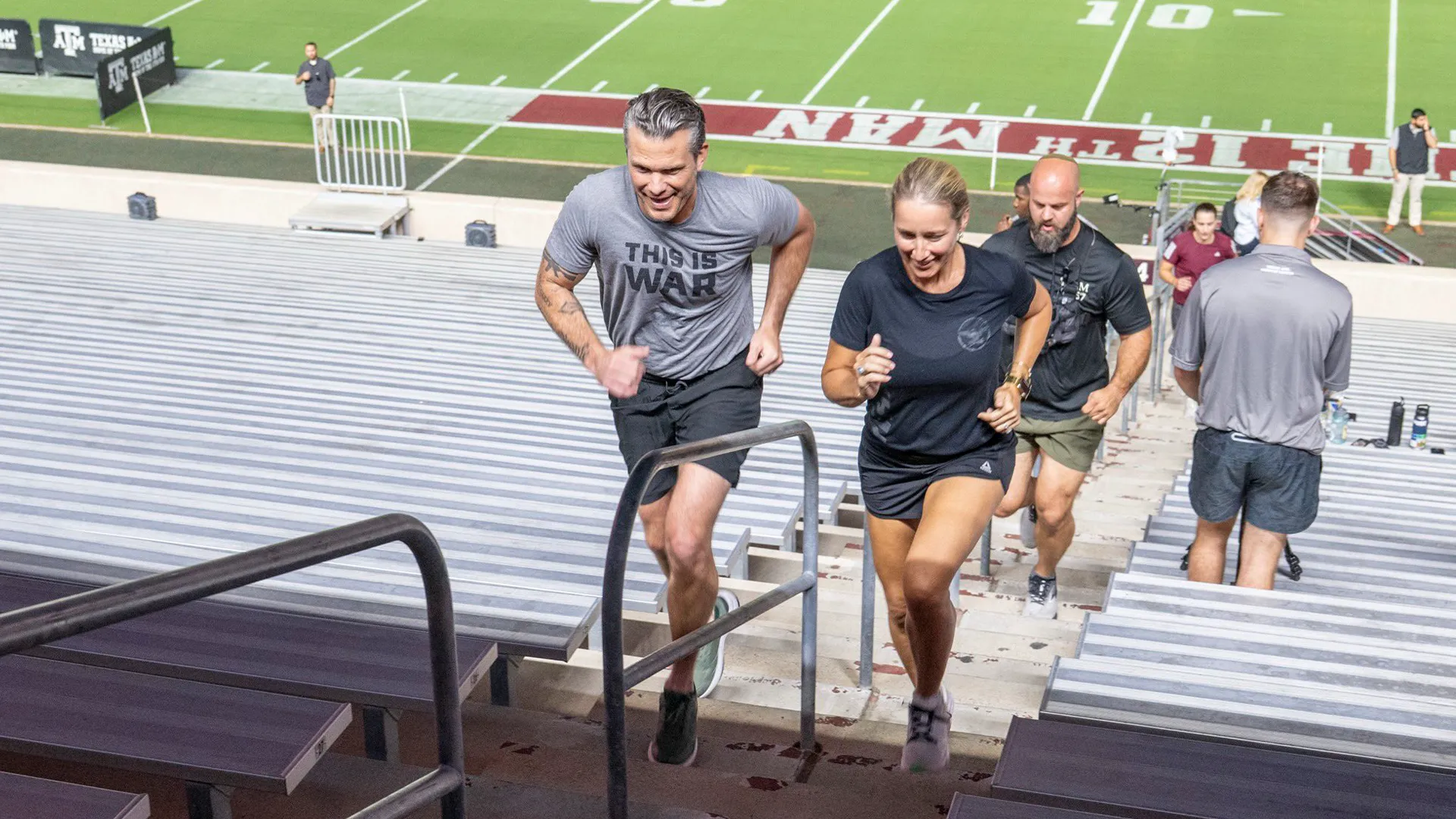 People climb stadium bleachers during a fitness activity near a football field.