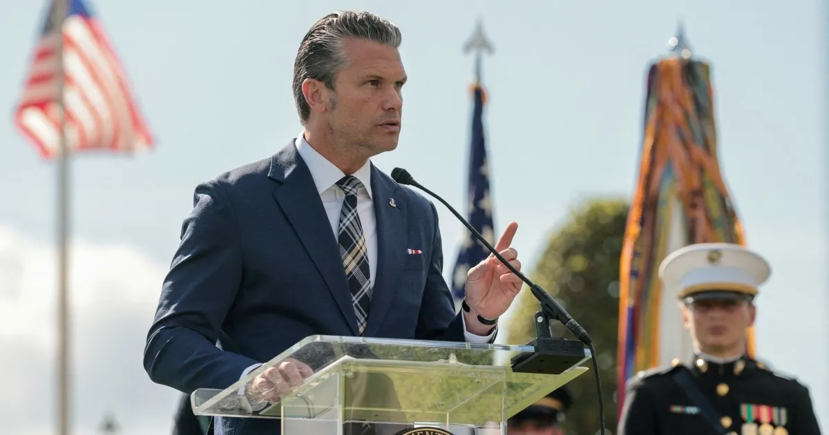 A man in a navy suit speaks at a clear podium beside a uniformed Marine and flags.