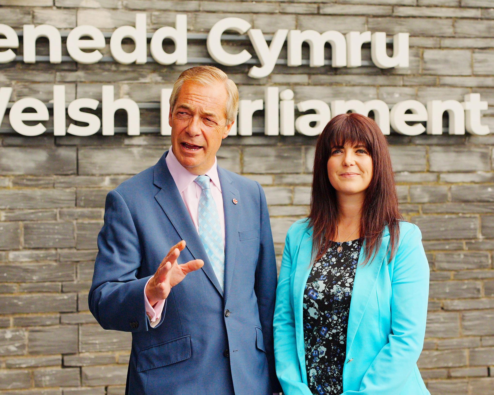 Man in blue suit and woman in turquoise jacket standing before a Welsh-language sign