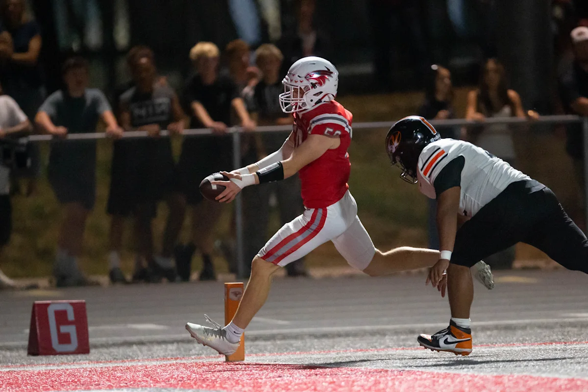 Football player in red runs with the ball as a defender in white pursues him.