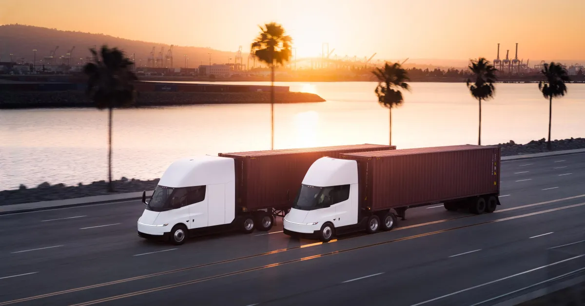 Two white semi trucks with dark trailers drive along a waterfront road at sunset.