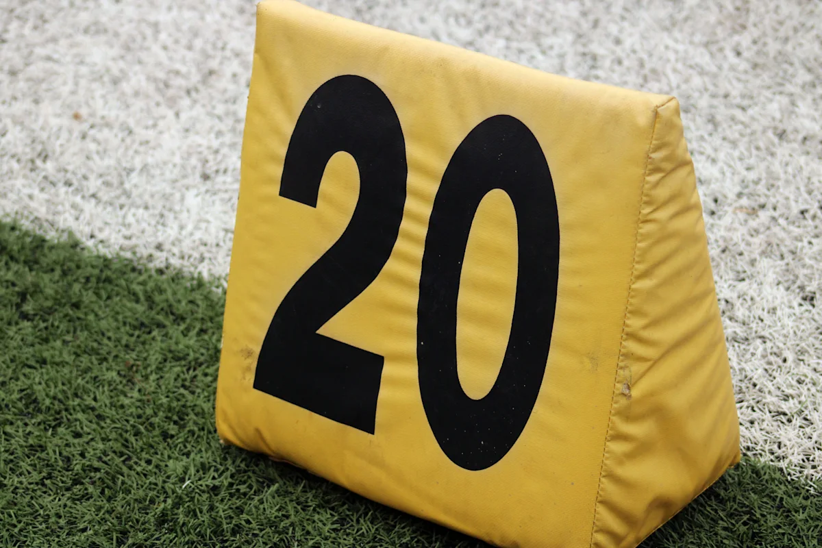 Yellow football yard marker displaying number 20 on artificial turf near a white sideline.