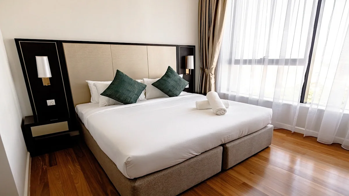 Bright hotel bedroom with white bed, green pillows, rolled towels, wood flooring, and large windows.
