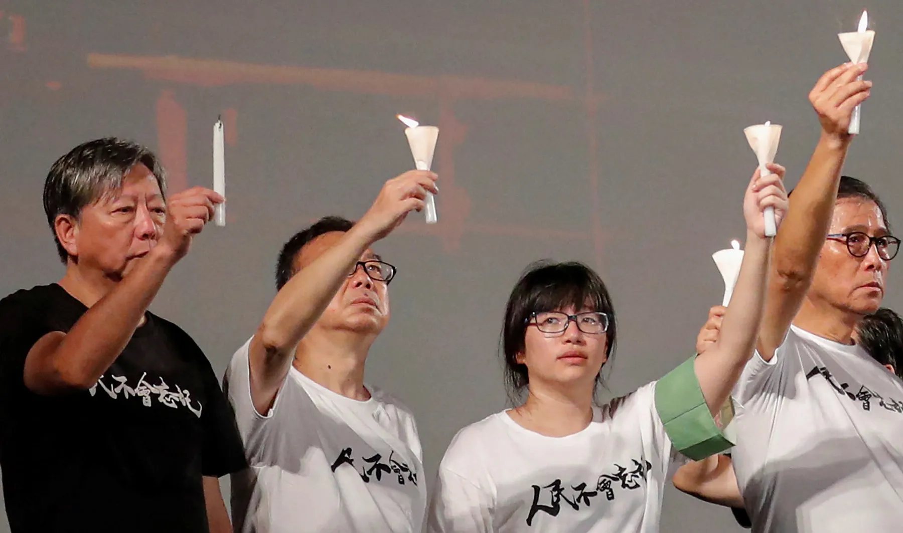 Five people raise candles during a solemn gathering, with several wearing shirts bearing Chinese characters.