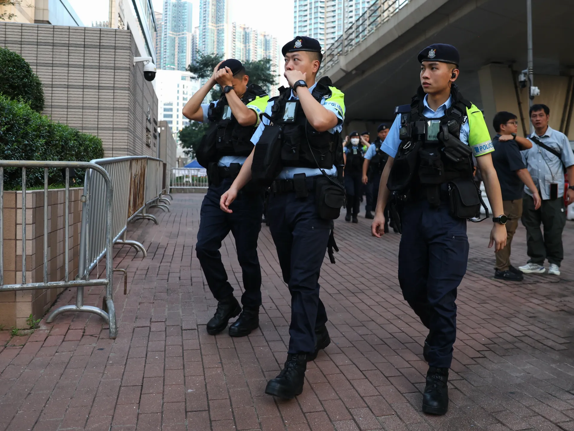 Uniformed police officers walk along a city sidewalk beside metal barriers and several bystanders.