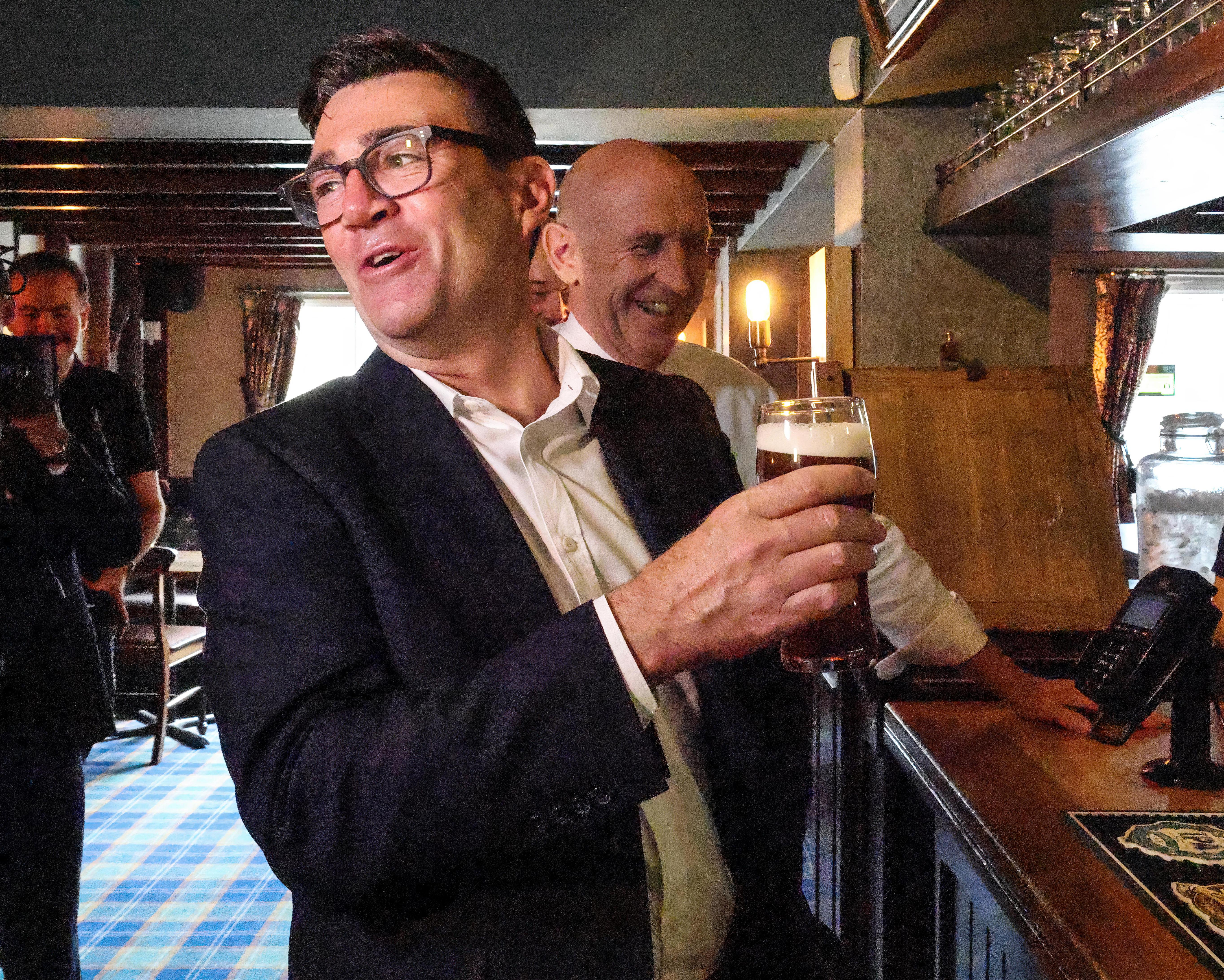 Man in glasses and suit holds a pint in a warmly lit bar beside another man