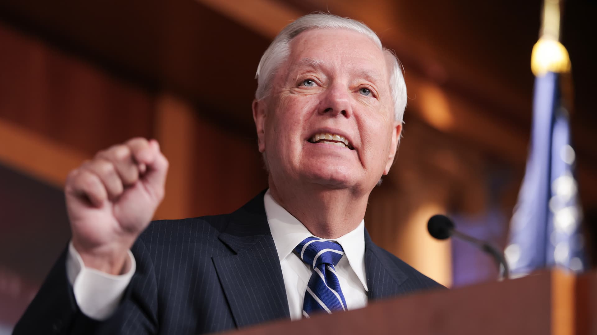 White-haired man in suit raises a fist while speaking at a podium near a U.S. flag