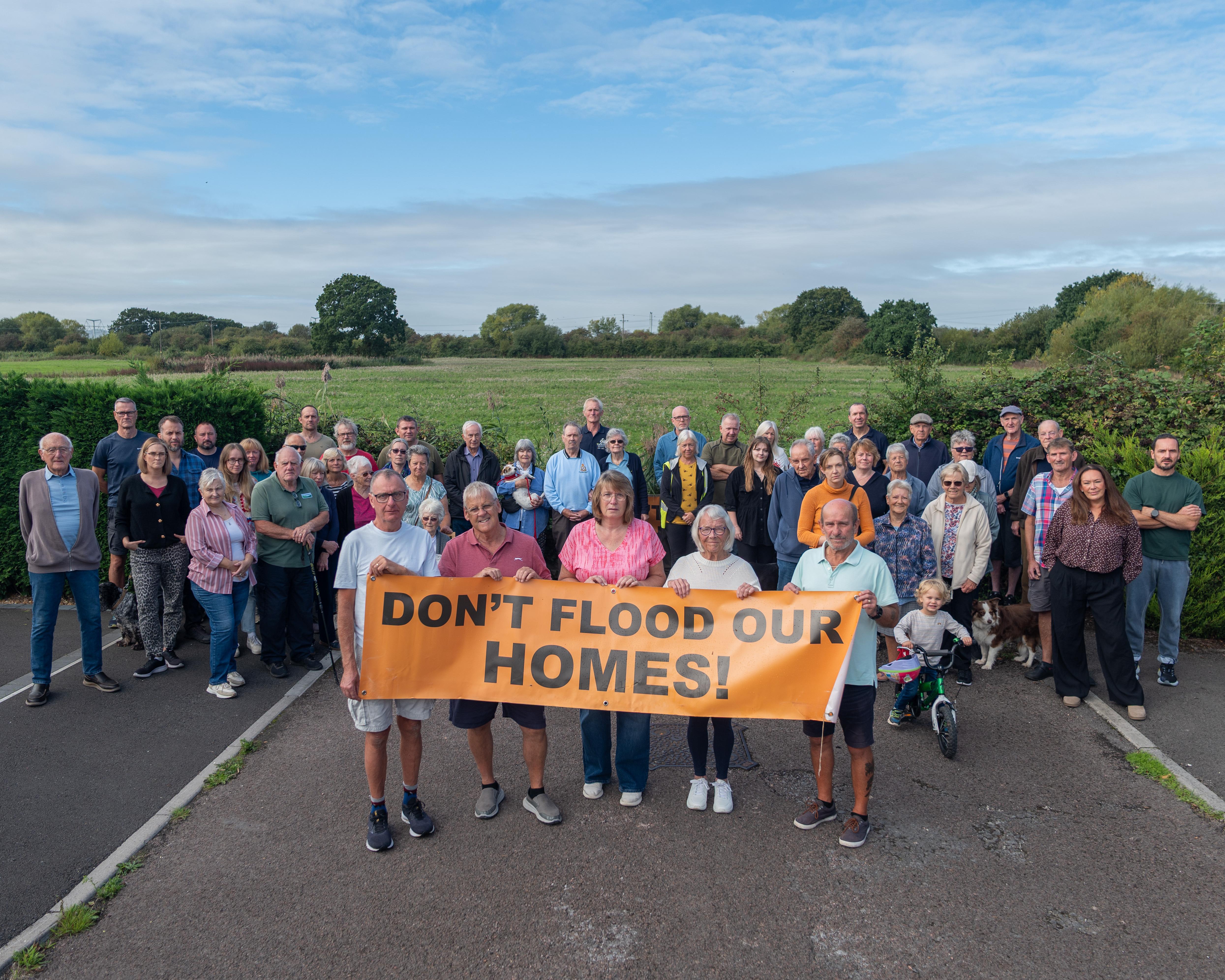 A large group poses behind an orange banner reading “Don’t flood our homes!” beside a field.
