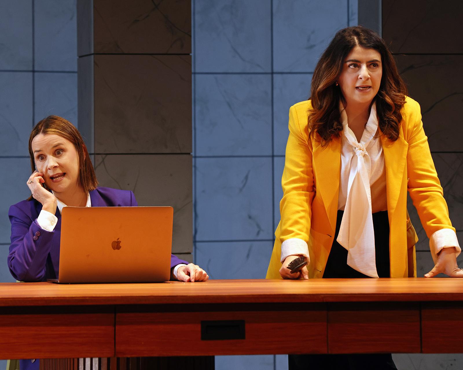 Two women in colorful jackets appear behind a desk, one using a laptop and the other holding a brush.