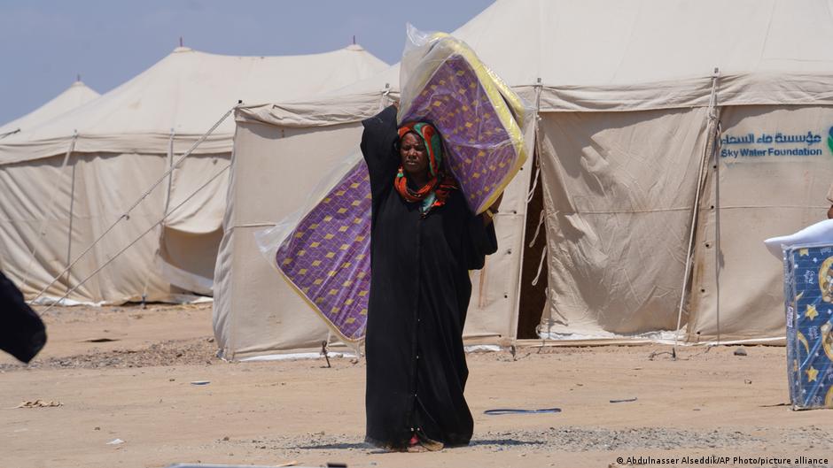A woman in a black abaya stands before large white tents, carrying a purple patterned bundle.