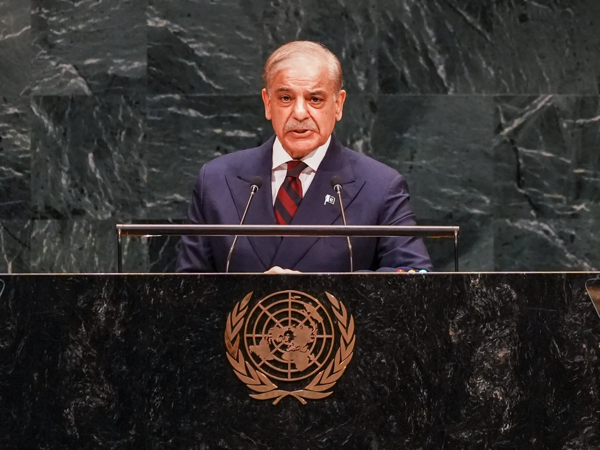 A suited man speaks at a United Nations podium, with the organization’s emblem visible below.