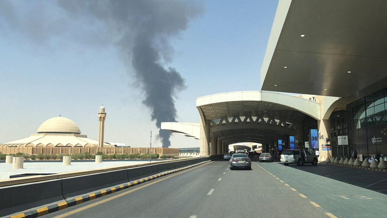 Road beside an airport terminal and mosque, with a thick column of dark smoke rising in the distance.