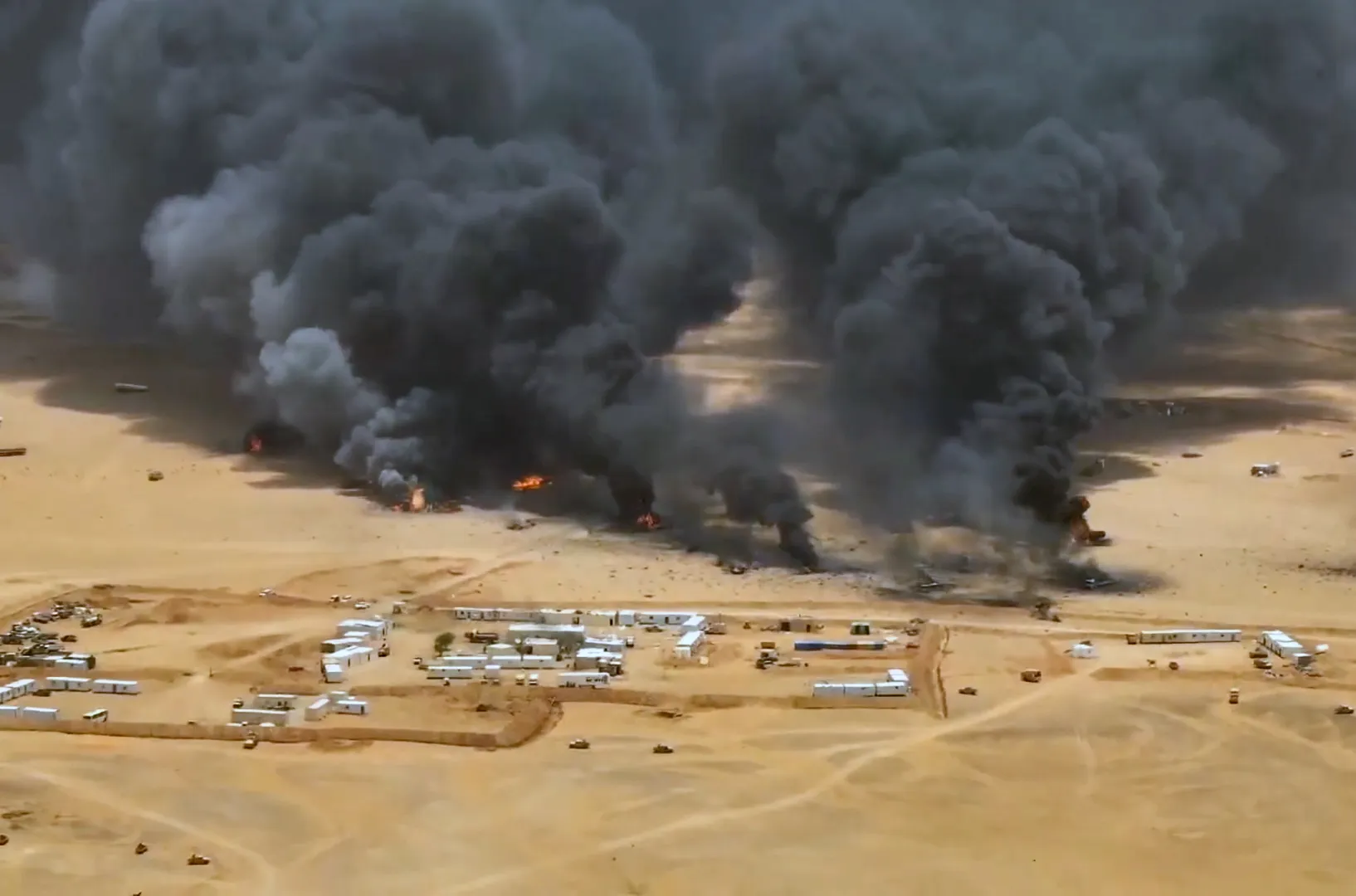 Aerial view of thick black smoke and flames rising from a sandy compound in the desert.