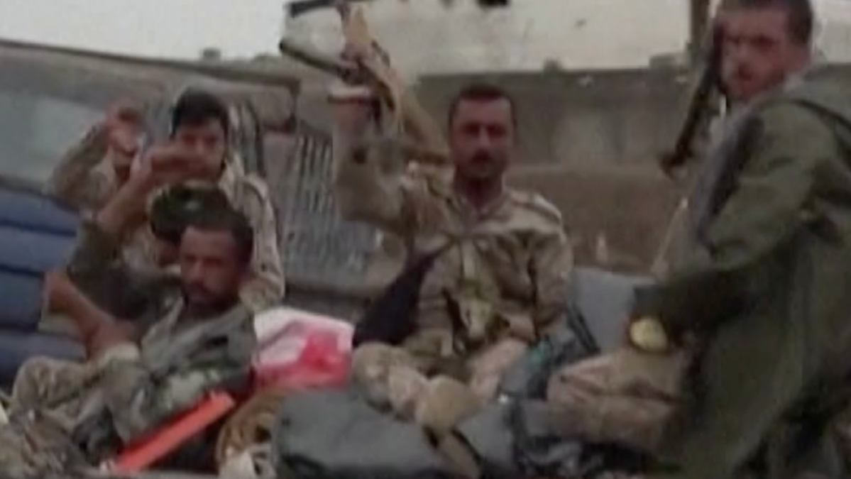 Several men in military-style clothing sit and stand in the back of a vehicle.