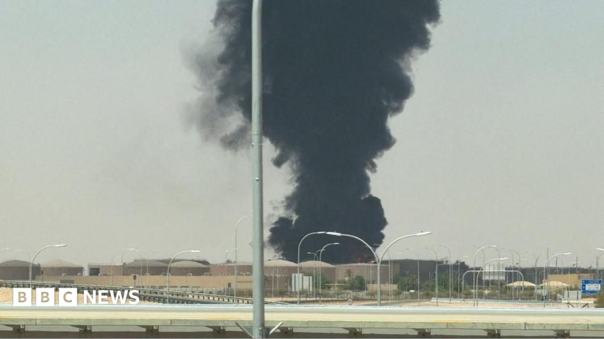 Thick black smoke rises behind buildings near a multilane roadway under a hazy sky.