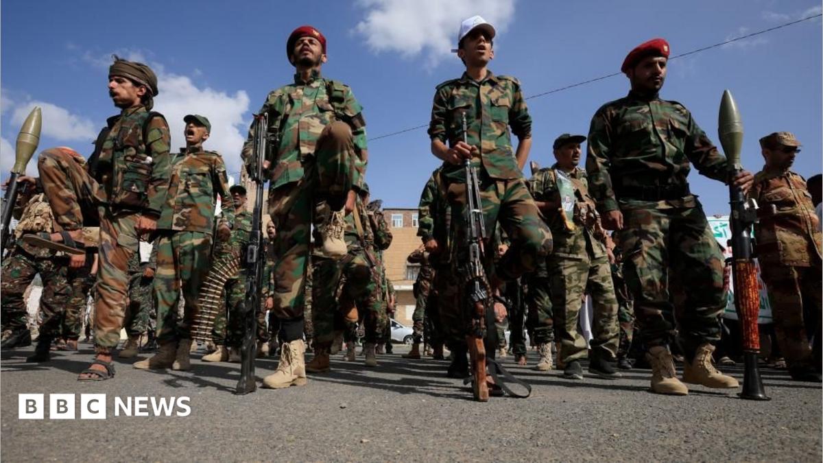Armed men in camouflage uniforms march along a city street beneath a blue sky.