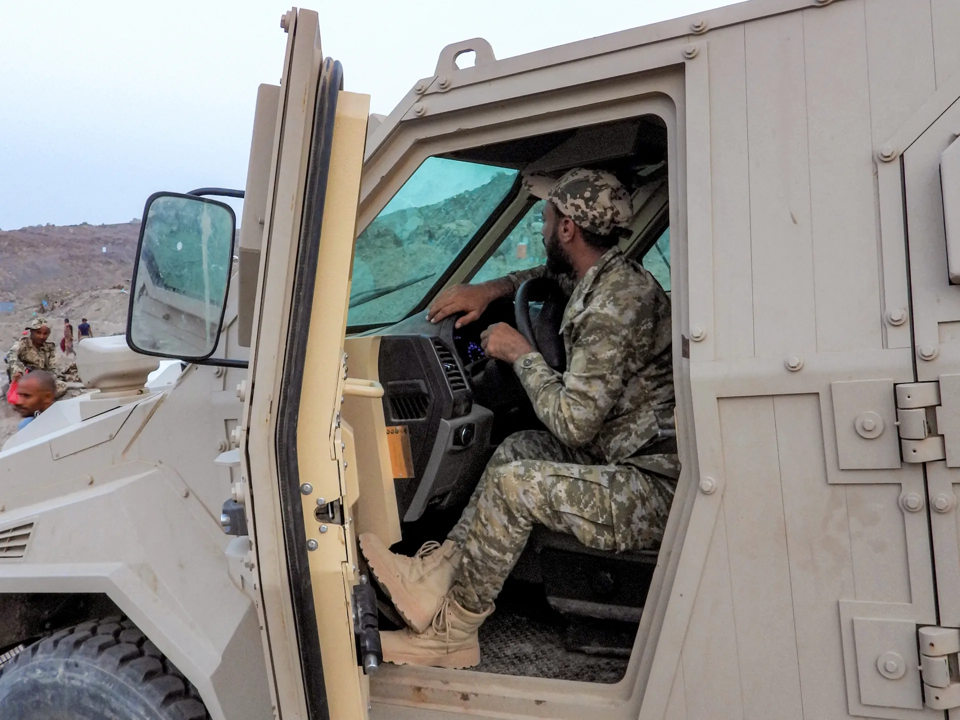 Soldier in camouflage sits inside an armored vehicle near a rocky hillside.